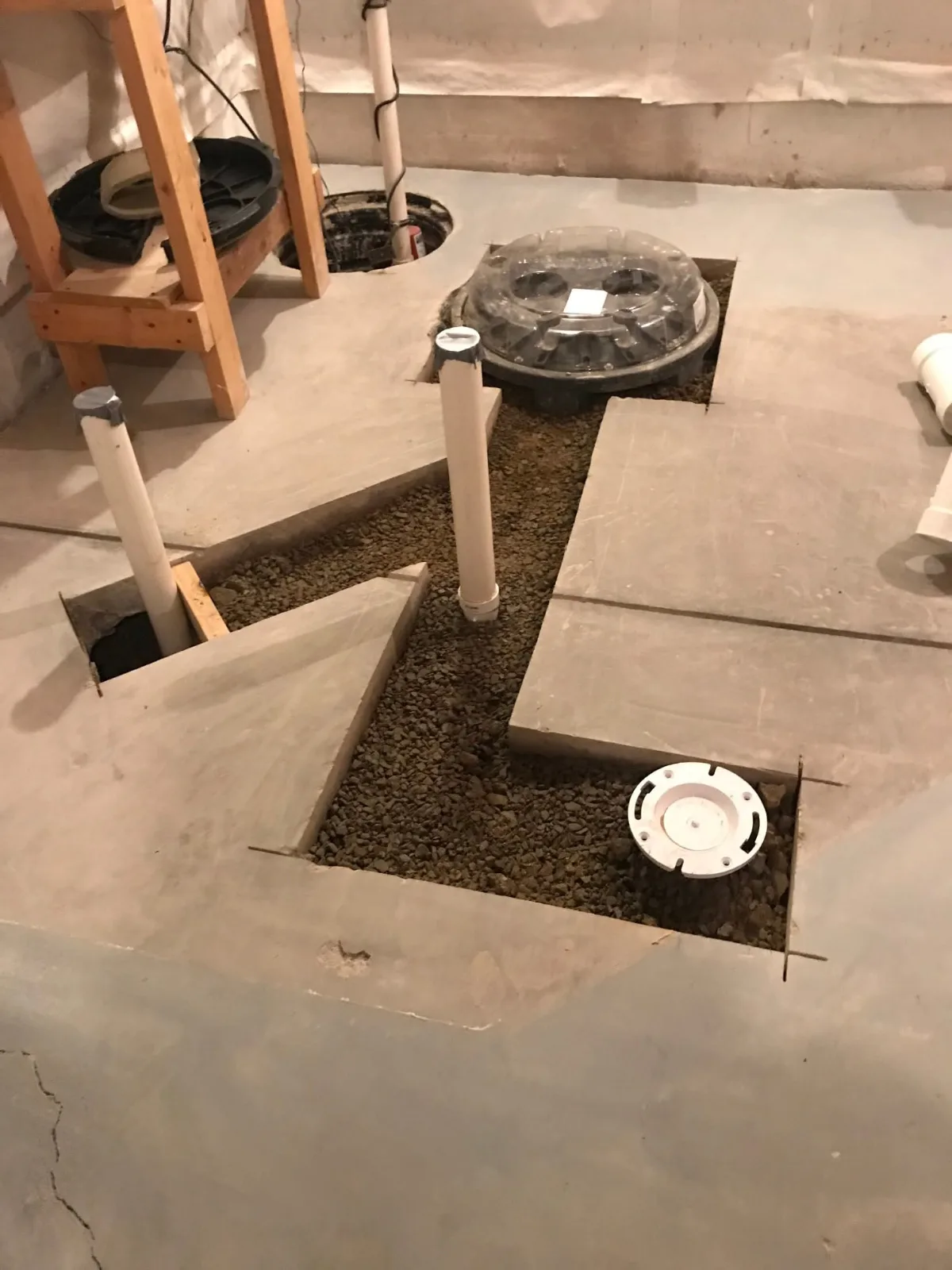 A basement floor cut open for a new bathroom: drain lines in gravel, a toilet flange and an ejector pit