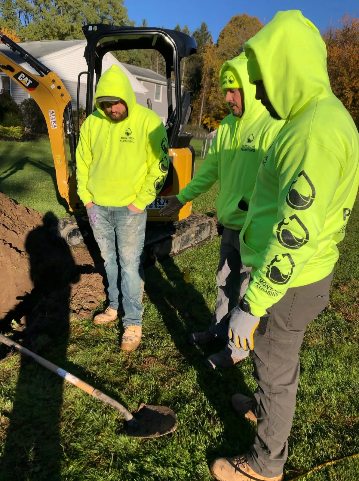Three of the Montoya Plumbing crew in matching hi-vis hoodies beside a mini excavator, looking into a dig