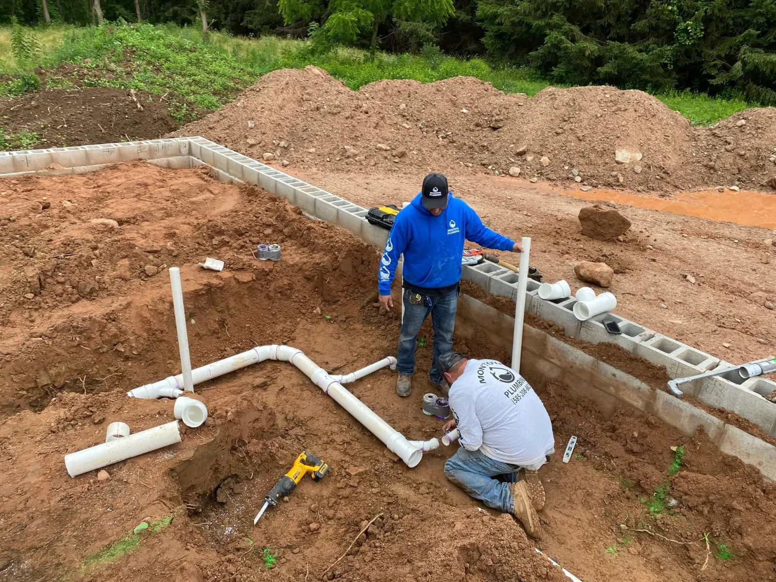 Two Montoya Plumbing plumbers fitting white PVC drain lines inside a new block foundation