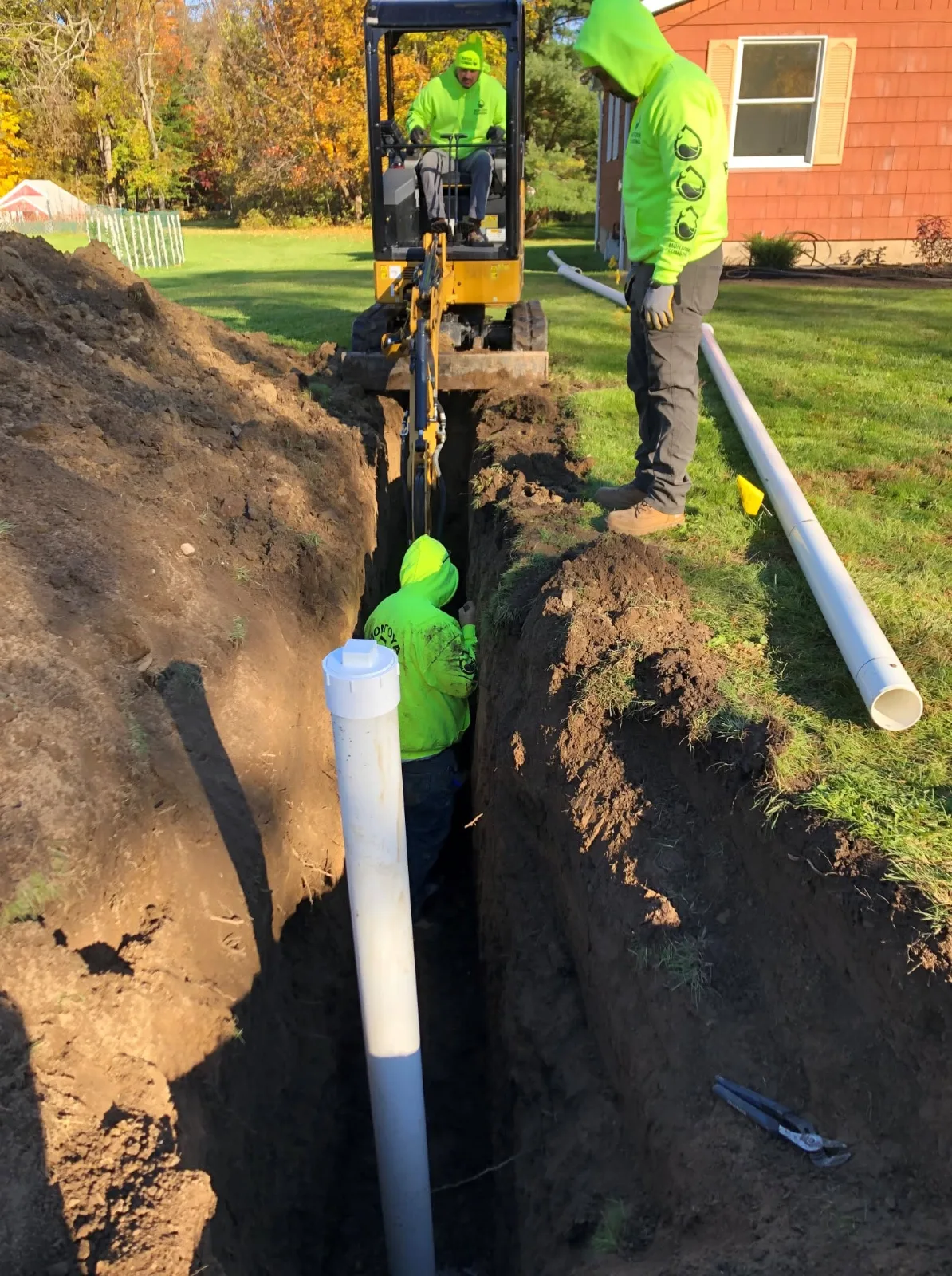The crew digging a sewer line trench with a mini excavator, one of them down in the trench