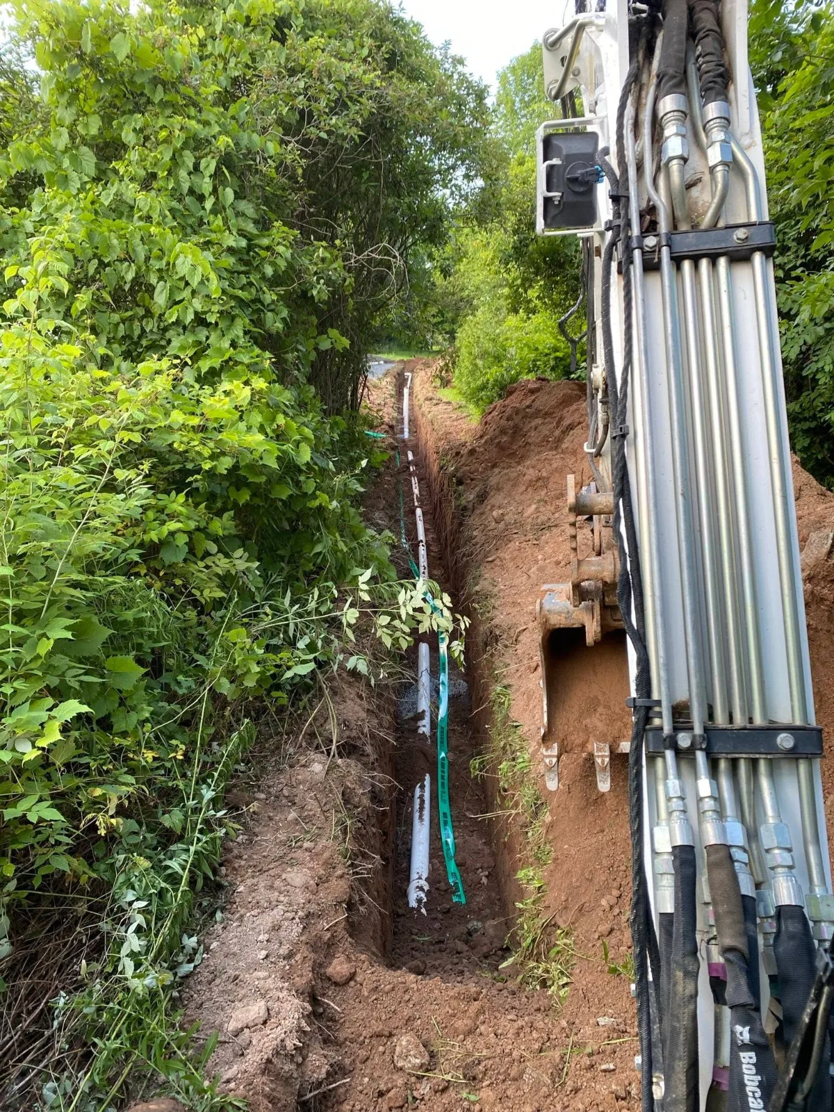 A long trench with new white pipe and green warning tape, beside an excavator arm